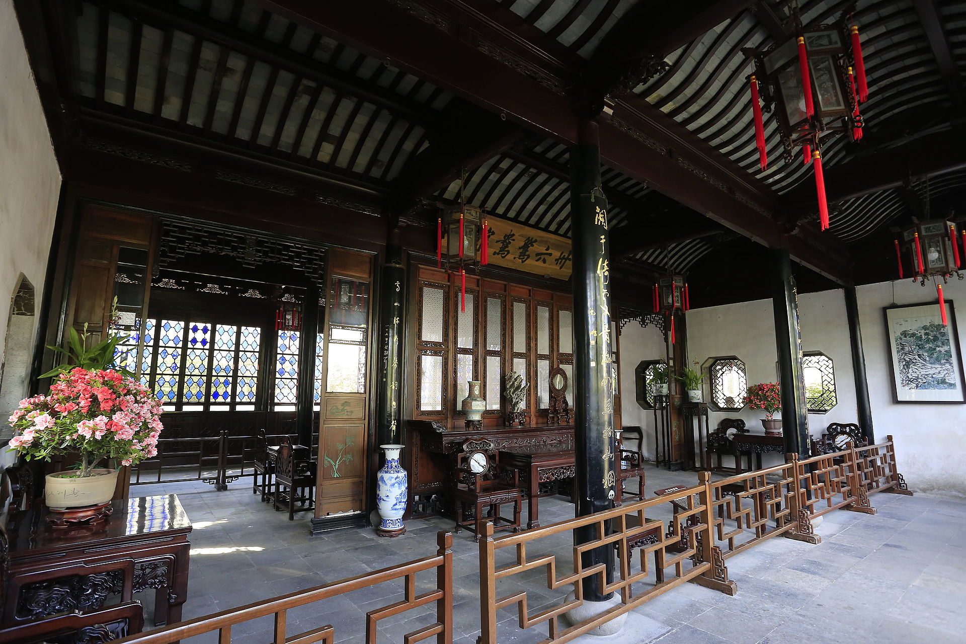 Interior of a Chinese pavilion inside; black columns with gold Chinese calligraphy, wooden sign with Chinese calligraphy in black, ornate wooden fence and furniture, lanterns hanging from ceiling, stained glass windows, vases.
